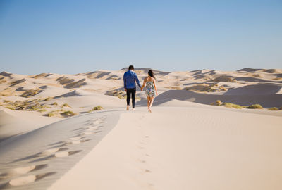 Rear view of couple walking on sand at desert against sky