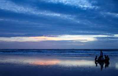 Silhouette people riding on beach against sky during sunset