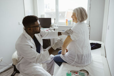 Young male healthcare worker vaccinating senior female patient sitting on bed at healthcare center