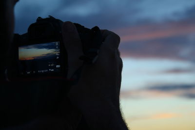 Close-up of hand holding camera against sky during sunset