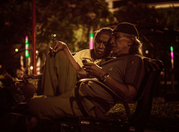 Man using mobile phone by woman while sitting on chair at night