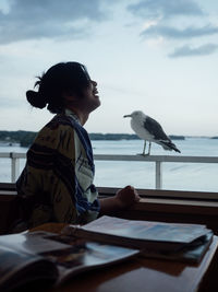 Cheerful woman sitting by seagull perching on railing