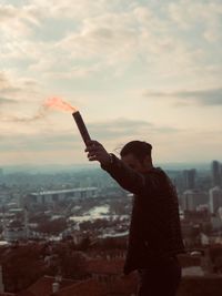 Side view of man holding distress flare while standing in city against sky during sunset