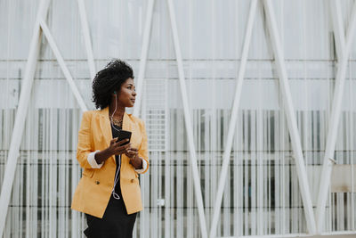 Businesswoman listening music through earphones while looking while standing against building