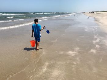 Rear view of boy with bucket walking on beach against sky