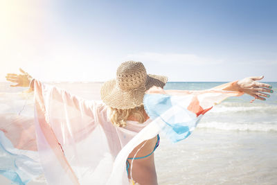 Rear view of young woman with arms outstretched standing at beach against sky