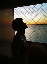 Silhouette man standing by fence against sky during sunset