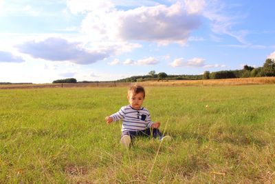Portrait of cute baby boy sitting on grassy field against sky