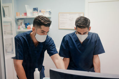 Anonymous young male doctors in respiratory masks and uniforms working on desktop computer in clinic