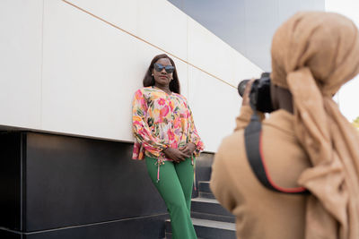Back view of unrecognizable black muslim female photographer in casual clothes and hijab taking photos of confident friend standing near modern building on stairs in city