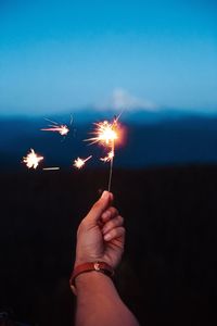 Cropped image of hand holding sparkler on field against sky at dusk