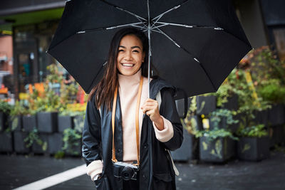 Smiling woman with camera carrying umbrella while standing in city