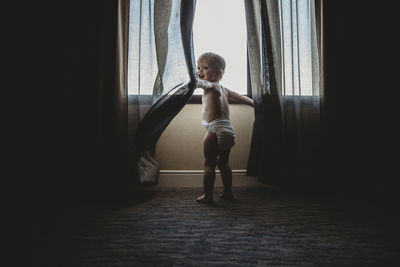 Rear view of shirtless baby boy playing with curtains while standing by window at home