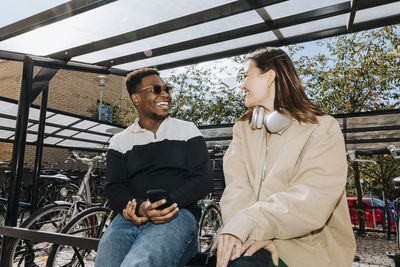 Happy multiracial male and female friends talking while sitting on railing at bicycle parking station