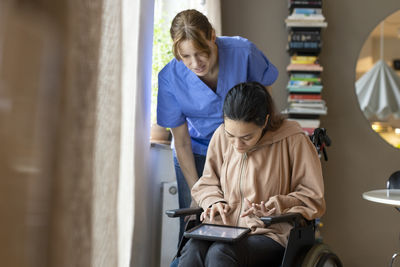 Female caregiver assisting woman using tablet pc while sitting on wheelchair at home
