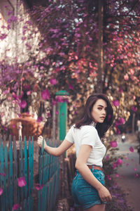 Portrait of young woman standing by fence against trees at park