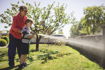 Full length of grandmother assisting grandson in spraying water on surrounding wall at yard