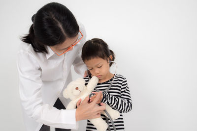 Doctor with girl holding stethoscope on teddy bear while standing against white background