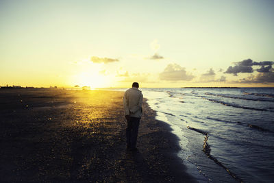 Rear view of man standing on beach against sky during sunset