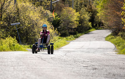 Girl riding pedal cart on road in distance.