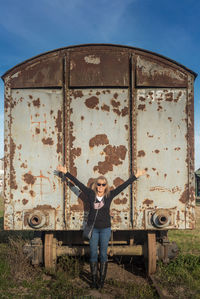 Adult woman wearing sunglasses smiling with her arms raised and abandoned train carriage