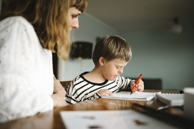 Boy with down syndrome writing in book while sitting by mother at table