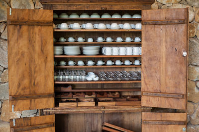 Crockery arranged on shelves of wooden cupboard against stone wall at home