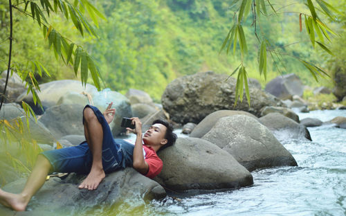 Full length of man relaxing on rock while using smart phone by river