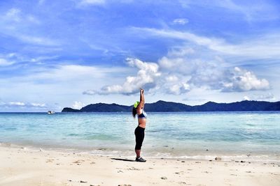 Full length of woman exercising at beach against cloudy sky on sunny day