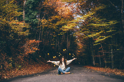 Positive curly haired woman in sweater throwing dry leaves in picturesque autumn forest 