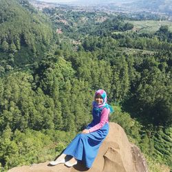 High angle portrait of smiling woman sitting on rock at mountain peak against landscape