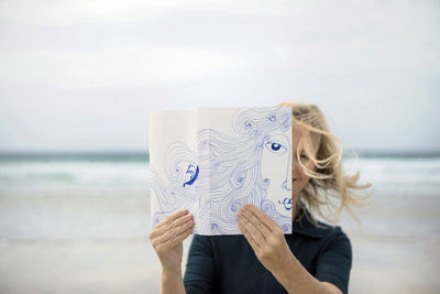 Woman covering face with book, reading poetry on beach