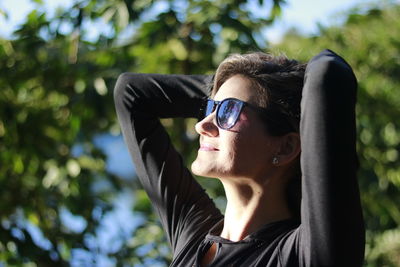 Woman with hands behind head wearing sunglasses against trees