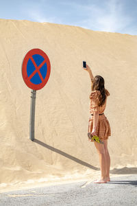 A image of a female model taking photograph with mobile phone of a sign post buried in sand