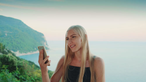 Portrait of smiling young woman using smart phone against sea