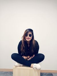 Portrait of young woman wearing sunglasses while sitting against wall