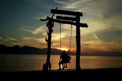 Silhouette people sitting on swing at beach against sky during sunset