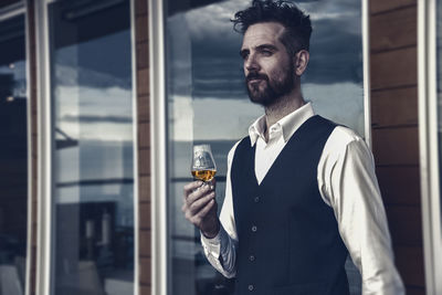 Confident man holding drink while standing against window