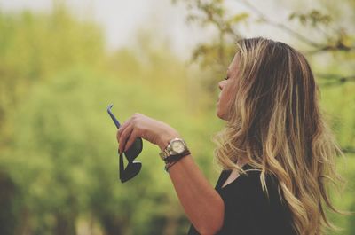 Side view of woman holding sunglasses while standing against trees