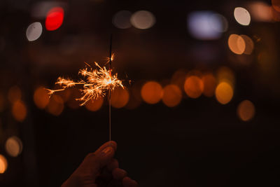 Cropped hand holding burning sparkler against illuminated lights at night