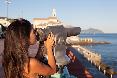 Side view of young woman looking at sea through coin-operated binoculars in city