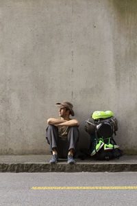 Man sitting with backpack on footpath against wall