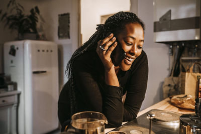 Young woman talking through smart phone while leaning on kitchen counter