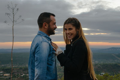 Side view of young couple standing against sky during sunset