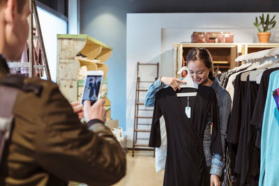 Man photographing smiling woman on smart phone holding dress at store