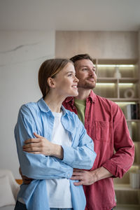 Gentle husband and tender wife standing embraced, looking at window, thinking of family, dreaming