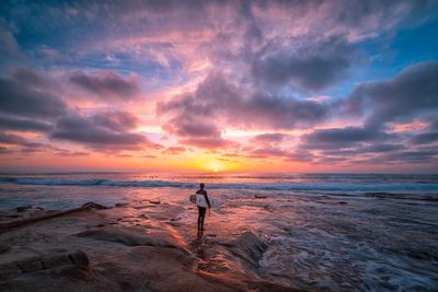 Rear view of man with surfboard standing at beach against cloudy sky during sunset