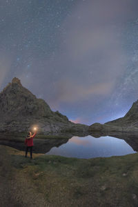 Back view of unrecognizable male tourist with torch admiring snowy mounts reflecting in water under starry sky at night