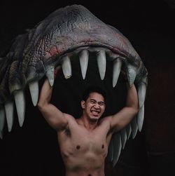 Shirtless man standing below dinosaur statue against black background