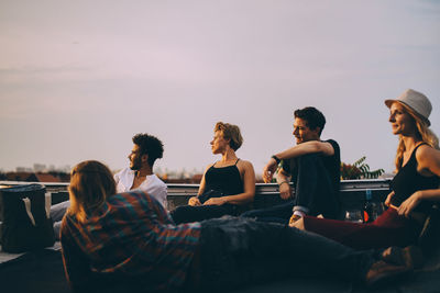 Friends looking away while relaxing together during rooftop party against sky in city